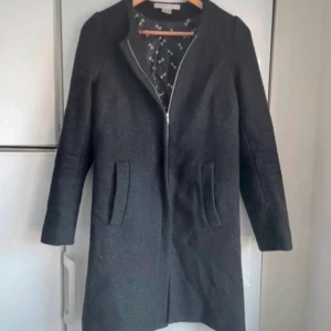 Mörkgrå ullkappa från Filippa K - Stilren mörkgrå ullkappa från Filippa K med rak passform och diskret dragkedja framtill. Kappan har två fickor och ett svart innerfoder med mönster. Perfekt för kyliga dagar och ger en klassisk look.