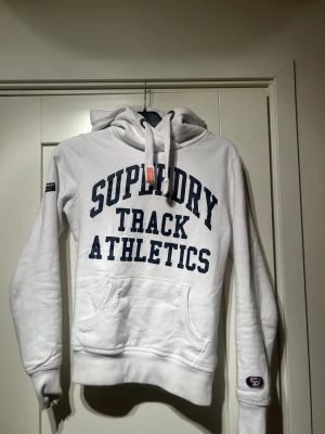 Vit Superdry hoodie med tryck - Snygg vit hoodie från Superdry med mörkblå text 'Superdry Track Athletics' på framsidan och siffran 72 på ryggen. Klassisk känguruficka, dragsko i huvan och ribbade muddar. Perfekt för en sportig och avslappnad stil. Tillverkas inte längre 
