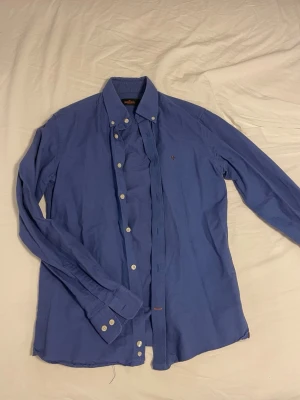 Blå skjorta från Morris med brodyr - Säljer en klassisk blå skjorta från Morris med button down-krage och guldfärgad/brun broderad logga på bröstet. Storlek S