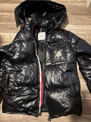 Svart dunjacka från Moncler - Snygg svart dunjacka från Moncler med glansig finish, huva och dragkedja framtill. Jackan har en bröstficka med Moncler-logga och tryckknapp samt två sidofickor. Perfekt för kalla vinterdagar och riktigt trendig look. Kan gå ner i pris vid snabb affär 