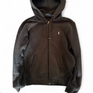 Svart hoodie från Polo Ralph Lauren - Klassisk svart hoodie från Polo Ralph Lauren med dragkedja framtill, två fickor och justerbar huva. Ikoniska lilla loggan broderad på bröstet. Tillverkad i mjukt bomullsmaterial som är skönt att ha på sig. Perfekt för en avslappnad och stilren look.