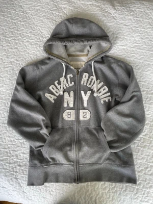 vintage Grå hoodie från Abercrombie & Fitch 2000s - Står XL men sitter som en verklig M!! Snygg grå hoodie från Abercrombie & Fitch med dragkedja, huva och vita broderade bokstäver på bröstet. Modellen har kängurufickor och vita snören i huvan. Mjuk insida i frottéliknande material och detaljer i beige vid huvkanten.