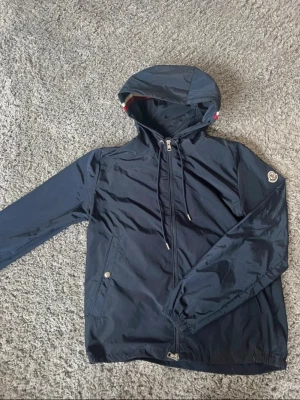 Mörkblå vindjacka från Moncler - Snygg mörkblå vindjacka från Moncler med huva och dragkedja framtill. Jackan har två fickor med tryckknappar och justerbar dragsko i huvan. Moncler-logga på ärmen och diskreta röda/vita detaljer i huvan. Perfekt för blåsiga dagar.
