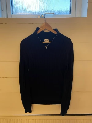 Mörkblå stickad tröja John Henric - Snygg mörkblå stickad tröja från John Henric med halv dragkedja och hög krage. Tröjan har ett klassiskt kabelstickat mönster och långa ärmar. Perfekt för lager på lager och en stilren look.