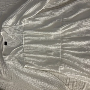 Vit långärmad klänning Gina Tricot - Säljer en vit långärmad klänning från Gina Tricot med v-ringad hals och strukturerat tyg. Klänningen har en luftig passform och är perfekt för dig som gillar en enkel men stilren look. Materialet känns mjukt och bekvämt mot huden.