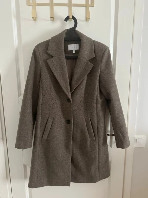 Brun kappa från VILA - Stilren brun kappa från VILA med klassisk krage och två knappar framtill. Kappan har två snedställda fickor och en rak passform som ger en tidlös look. Perfekt för kyliga dagar och enkel att matcha med olika outfits.