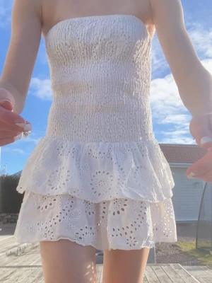Vit axelbandslös kort klänning - Söt vit axelbandslös klänning med smockad överdel och dubbla volanglager i kjolen. Klänningen har broderade hålmönster och ger en luftig och somrig känsla. Perfekt för varma dagar och enkel att styla med accessoarer.