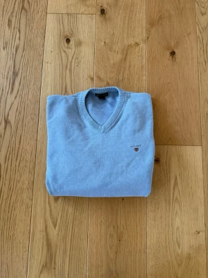 Ljusblå V-ringad tröja från Gant - Snygg ljusblå V-ringad tröja från Gant med diskret logga broderad på bröstet. Tröjan har långa ärmar, ribbade muddar och är tillverkad i mjukt material som känns skönt mot huden. Perfekt för en clean och stilren look.