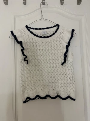 Vit stickad topp med mörkblå kant Lindex - Söt vit stickad topp från Lindex med mörkblå vågig kant runt hals, ärmar och nederkant. Toppen är ärmlös och har ett fint hålmönster i tyget. Perfekt för en fräsch och somrig look.