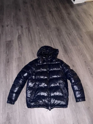 Mörkblå dunjacka från Moncler - Snygg mörkblå dunjacka från Moncler med glansig finish och huva. Jackan har två sidofickor med dragkedja och en Moncler-logga på ärmen. Perfekt för kalla vinterdagar tack vare det tjocka dunfodret och den klassiska puffer-modellen.
