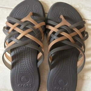 Bruna och grå Crocs sandaler - Säljer ett par snygga sandaler från Crocs med flätade remmar i brunt och grått. Sulan är mörkgrå och har en bekväm, lätt vadderad struktur. Perfekta för sommaren med sin öppna design och flexibla material. Stl:34