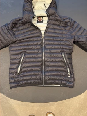 Svart dunjacka från Colmar Originals - Snygg svart dunjacka från Colmar Originals med horisontella sömmar, två dragkedjeförsedda fickor och huva. Jackan har en lätt glansig finish och ljusgrått innerfoder. Perfekt för våren som är här 