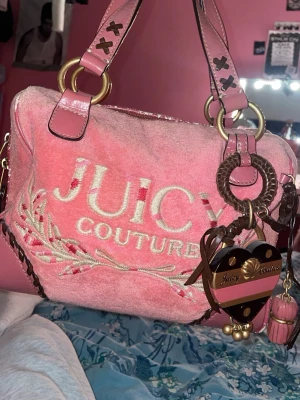 Juicy Couture rosa handväska - Säljer en ikonisk rosa handväska från Juicy Couture i en vacker rosa/korallfärg med läderdetaljer och guldiga metalldelar. Väskan har broderad logga framtill, hjärtformad detalj bak och söta hängen i form av hjärta och tofs. Perfekt för dig som älskar Y2K-stil och vill sticka ut. Mindre slitningar på insida och utsida vilka syns på bilderna. Perfekt storlek, rymmer kläder, böcker plånbok mm.