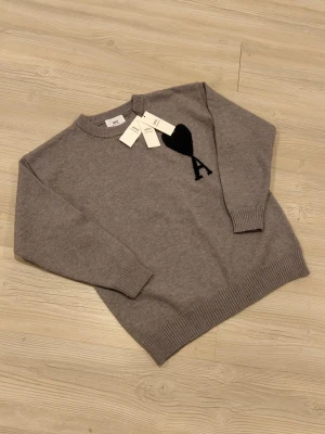 Grå stickad tröja från Ami Paris - Säljer en grå stickad tröja från Ami Paris med svart hjärta och bokstaven A på bröstet. Tröjan har rund hals, ribbade muddar och är tillverkad av ekologisk bomull. Perfekt för dig som gillar stilrena och trendiga plagg.