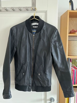 Svart skinnjacka These Glory Days - Snygg svart skinnjacka från These Glory Days i klassisk bombermodell. Jackan har ribbade muddar vid krage, ärmslut och nederkant samt två snedställda fickor med knapp. Fodret är rutigt i blått och vitt. Perfekt för dig som gillar stilren och tidlös look. Den har två innerfickor med dragkedja, en på varje sida.                                         Den är använd men är fortfarande i svinbra skick och är av 100% skinn🖤
