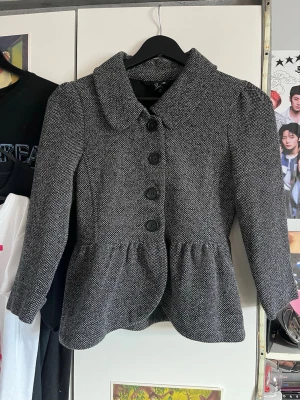 Grå kappa med knappar - Stilren grå kappa med markerad midja och stora svarta knappar framtill. Kappan har en bred krage och lätt puffade axlar för en trendig siluett. Materialet är strukturerat och ger en klassisk look. Perfekt för dig som vill ha en snygg och tidlös jacka. Trekvartsärmar!