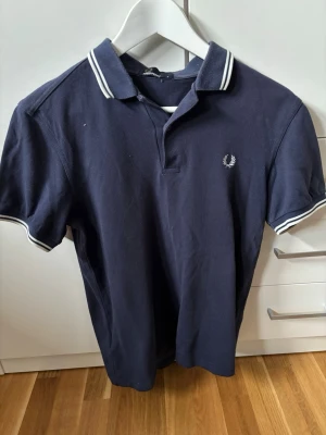 Marinblå Fred Perry piké t-shirt - Klassisk marinblå piké t-shirt från Fred Perry med vit logga broderad på bröstet och vita ränder på krage och ärmslut. Tillverkad i bomull och har en normal passform med korta ärmar och krage.