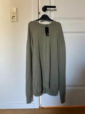 Olivgrön stickad tröja med rund hals (Ny med prislapp) - Enkel och stilren olivgrön stickad tröja med rund halsringning och långa ärmar. Tröjan har ribbstickade muddar vid ärmslut och nederkant. Perfekt för lager-på-lager och passar till det mesta.