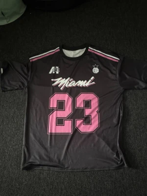 Svart Miami tröja med rosa tryck - Snygg svart sporttröja med texten 'Miami' i vitt och rosa samt stort nummer 23 i rosa framtill. Tröjan har vita och rosa detaljer på axlarna och en basketboll-logga på bröstet. Perfekt för basket eller streetwear.