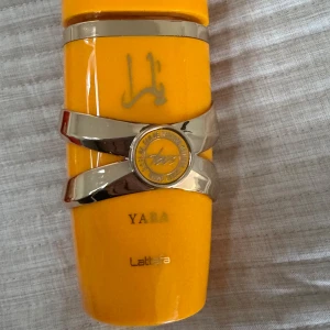 Gul parfymflaska Yara från Lattafa - Säljer en parfymflaska i klargul färg från Lattafa med namnet Yara. Flaskan har en unik form med silvriga detaljer och ett runt emblem framtill. Perfekt för dig som gillar snygga och färgstarka parfymflaskor.