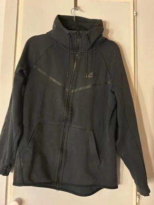 Svart Nike hoodie med dragkedja - Snygg svart hoodie från Nike med hel dragkedja framtill och två stora fickor. Hoodien har en hög krage, justerbar huva med snören och diskret Nike-logga på bröstet. Tillverkad i mjukt material som passar perfekt till en avslappnad streetwear-look.