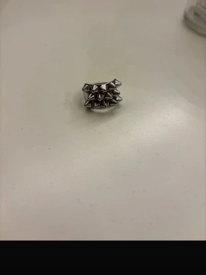 Silvrig ring med nitar - Cool silvrig ring med flera små pyramidformade nitar runt hela bandet. Ringen har en bred design och är tillverkad i metall, perfekt för dig som gillar en edgy och unik stil.