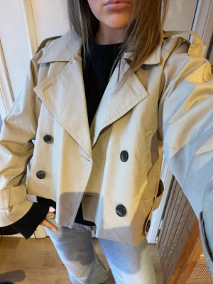 Croppad trenchcoat från Pull&Bear - Slutsåld! Stilren croppad trenchcoat från Pull&Bear i beige bomullsmix. Jackan har dubbelknäppning, bred krage och snygga detaljer med knappar vid ärmslut och axlar. Köptes på Zalando i  juli och har bara använts 4 gånger typ. Helt nytt skick! Kan ta fler bilder om önskat i annat ljus.