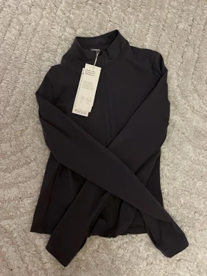 Grå träningsjacka från H&M Move - Snygg grå träningsjacka från H&M Move SoftMove-kollektionen. Jackan har hel dragkedja framtill, hög krage och är tillverkad i ett mjukt, stretchigt funktionsmaterial som andas. Perfekt för gymmet eller löprundan.