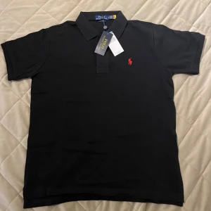 Svart pikétröja från Polo Ralph Lauren - Klassisk svart pikétröja från Polo Ralph Lauren med röd broderad logga på bröstet. Tröjan har krage, korta ärmar och knappar vid halsen. Perfekt för en stilren och tidlös look.