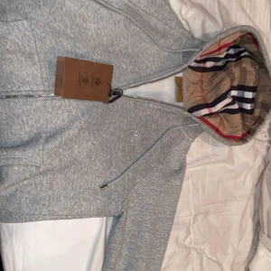 Grå hoodie från Burberry med huva - Snygg grå hoodie från Burberry med dragkedja och klassiskt rutigt foder i huvan. Märket är broderat på bröstet och huvan har justerbara snören. Perfekt för dig som gillar exklusiv streetwear och vill ha något stilrent men ändå lyxigt. Hör av er för minsta lilla fråga! Passar mig bra är 173cm.