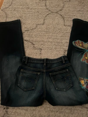 Mörkblå jeans med broderad detalj - Unika mörkblå jeans med superfina broderade fjärilar på ena benet. Jeansen är lågmidjade och är bootcut. Storlek S men passar mer som XS. Säljer för att de tyvärr är för små. Knappt använda och fint skick! 