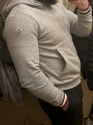 Grå Moncler hoodie med dragkedja - Snygg grå hoodie från Moncler med dragkedja framtill och klassiska Moncler-loggan på ärmen. Huva med dragsko och ribbade muddar med röd, vit och marinblå rand. Perfekt för en avslappnad och sportig stil. Tillverkad i mjukt bomullsmaterial.