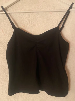 Svart ribbad linne - Säljer en svart ribbad linnetopp med smala axelband och lätt rynkning vid bysten. Perfekt att styla med jeans eller kjol för en enkel och snygg look.