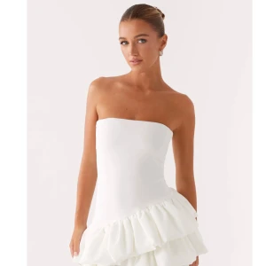 Vit axelbandslös volangklänning - Säljer en stilren vit axelbandslös kortklänning med volangdetaljer nedtill från Peppermayo. Klänningen har en figurnära passform upptill och är perfekt för sommarens alla tillfällen som tillexempel student. Materialet känns lätt och luftigt. Helt ny med prislapp kvar. 