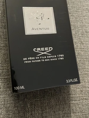 Creed Aventus Eau de Parfum 100ml - Lyxig parfym från Creed, Aventus, i en elegant svart förpackning med silverdetaljer och logotyp. Flaskan rymmer 100 ml och har en exklusiv design med en silverplatta framtill. En ikonisk doft som utstrålar stil och klass.