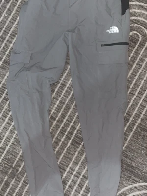 Grå cargobyxor från The North Face - Snygga grå cargobyxor från The North Face med flera praktiska fickor, inklusive en med dragkedja. Tillverkade i ett slitstarkt material som passar perfekt för en aktiv livsstil. Loggan syns tydligt på både benet och fickan. Raka ben och modern passform.