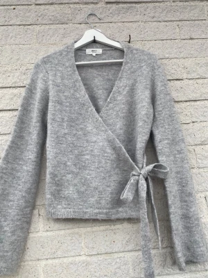 Grå omlottkofta från Gina Tricot - Mysig grå omlottkofta från Gina Tricot med knytband i midjan och lång ärm. Koftan är stickad i ett mjukt material och har en djup v-ringning framtill.   Knappst använd.