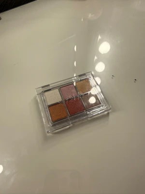 Ögonskuggspalett med sex nyanser - Säljer en ögonskuggspalett från Caia med sex skimrande nyanser i guld, koppar, rosa, champagne, brons och beige. Paletten har ett genomskinligt plastlock och fyrkantiga pans. Perfekt för dig som gillar att experimentera med olika färger och looks.