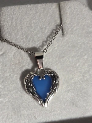 Halsband med blått lysande hjärta - Unikt halsband med ett hjärtformat hänge i blått som lyser i mörkret. Hänget är inramat av silverfärgade detaljer som liknar vingar. Kedjan är tunn och silverfärgad i rostfritt stål. Perfekt för dig som gillar smycken med en magisk touch.
