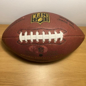 NFL amerikansk fotbollsläderboll - Säljer en klassisk amerikansk fotboll från NFL i brunt läder med vit snörning och tydlig NFL-logga i gult och svart. Perfekt för träning eller match med autentisk känsla och greppvänlig yta.