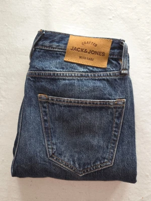 Blå jeans från Jack & Jones - Klassiska blå jeans från Jack & Jones i rak modell. Jeansen har en snygg mörkblå tvätt, fem fickor och kontrastsömmar. Tillverkade i slitstarkt denim med en läderpatch bak i midjan. Perfekta till vardagslooken.