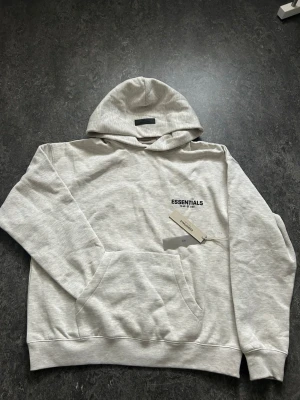 Grå Essentials hoodie - Snygg ljusgrå hoodie från Essentials med svart logga på bröstet och en grå patch på huvan. Klassisk passform med ribbade muddar och mjukt material, perfekt för en avslappnad streetwear-look. Dragsko saknas på huvan för en clean stil. Storlek L men passar bättre som XL eftersom essentials är oversized! Aldrig använt.