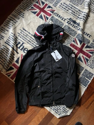 Svart vindjacka från Moncler - Size 2, Snygg svart vindjacka från Moncler med huva och dragkedja framtill. Jackan har Moncler-logga på ärmen och coola rödvita detaljer på huvan. Tillverkad i lätt, vindtätt material som passar perfekt för blåsiga dagar.