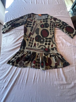 Mönstrad blus med volang - Unik kortklänning med abstrakt mönster i svart, beige och vinrött. Klänningen har trekvartsärm, rund halsringning och volang nedtill för en lekfull look. Perfekt för dig som gillar retrovibbar och vill sticka ut.