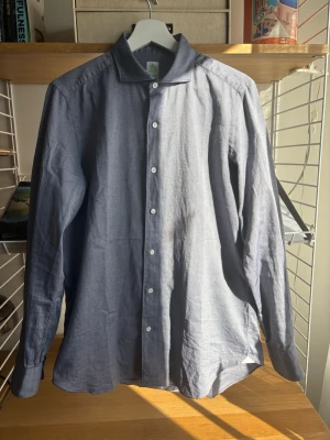 Finamore skjorta  - Klassisk blå skjorta i bomull med lång ärm och vita knappar hela vägen framtill. Skjortan har en enkel krage och rak passform, perfekt för en clean och stilren look. Materialet känns lätt och passar bra till både jeans och chinos. Storlek 40 vilket är en lite större M