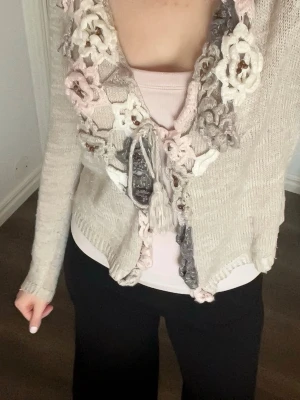 Beige kofta med virkade blommor - Unik beige kofta med långa ärmar och virkade blommor i vitt, rosa och grått längs kragen och öppningen. Koftan har knytband framtill och är tillverkad i ett mjukt, stickat material. Perfekt för dig som gillar romantiska detaljer och boho-stil.