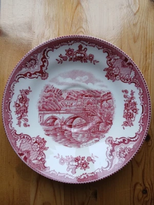 Rödvit tallrik Johnson Bros - Dekorativ tallrik från Johnson Bros med rödvit motiv av bro och landskap. Tallriken har detaljerade blommönster längs kanten och är tillverkad i porslin. Perfekt som samlarobjekt eller dekoration.