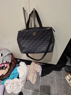 Svart handväska från Tiger of Sweden - Snygg svart handväska från Tiger of Sweden med monogrammönster och guldfärgade detaljer. Väskan har både handtag och en avtagbar axelrem, samt dragkedja upptill. Tillverkad i konstläder med stilren och modern design.