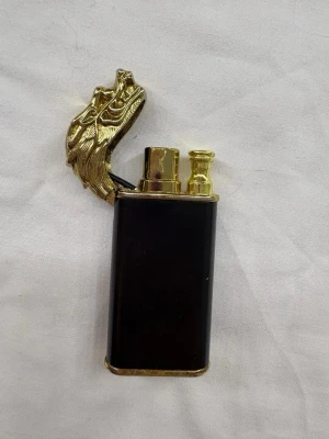 Tändare med drakhuvud i metall - Unik tändare med lock format som ett detaljerat drakhuvud i guldfärgad metall. Kroppen är svart och har guldfärgade detaljer. Perfekt för dig som gillar coola accessoarer med fantasytema.