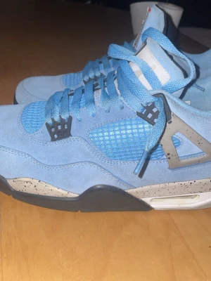 Jordan 4 University Blue sneakers - Snygga Jordan 4 University Blue sneakers i ljusblå mocka med meshpaneler och grå detaljer. Skorna har svart och grått stänkmönster på sulan, klassisk snörning och ikoniska plastdetaljer vid snörningen. Perfekta för dig som vill sticka ut med stil.